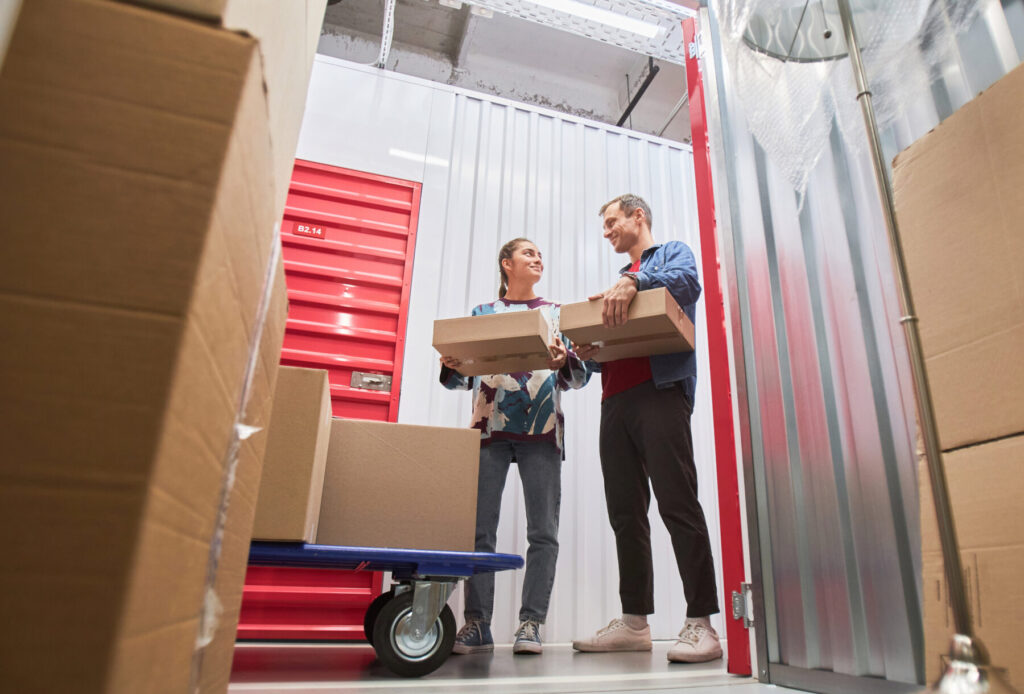 Two people organizing boxes in a self-storage unit with a red door and cardboard boxes in the foreground.