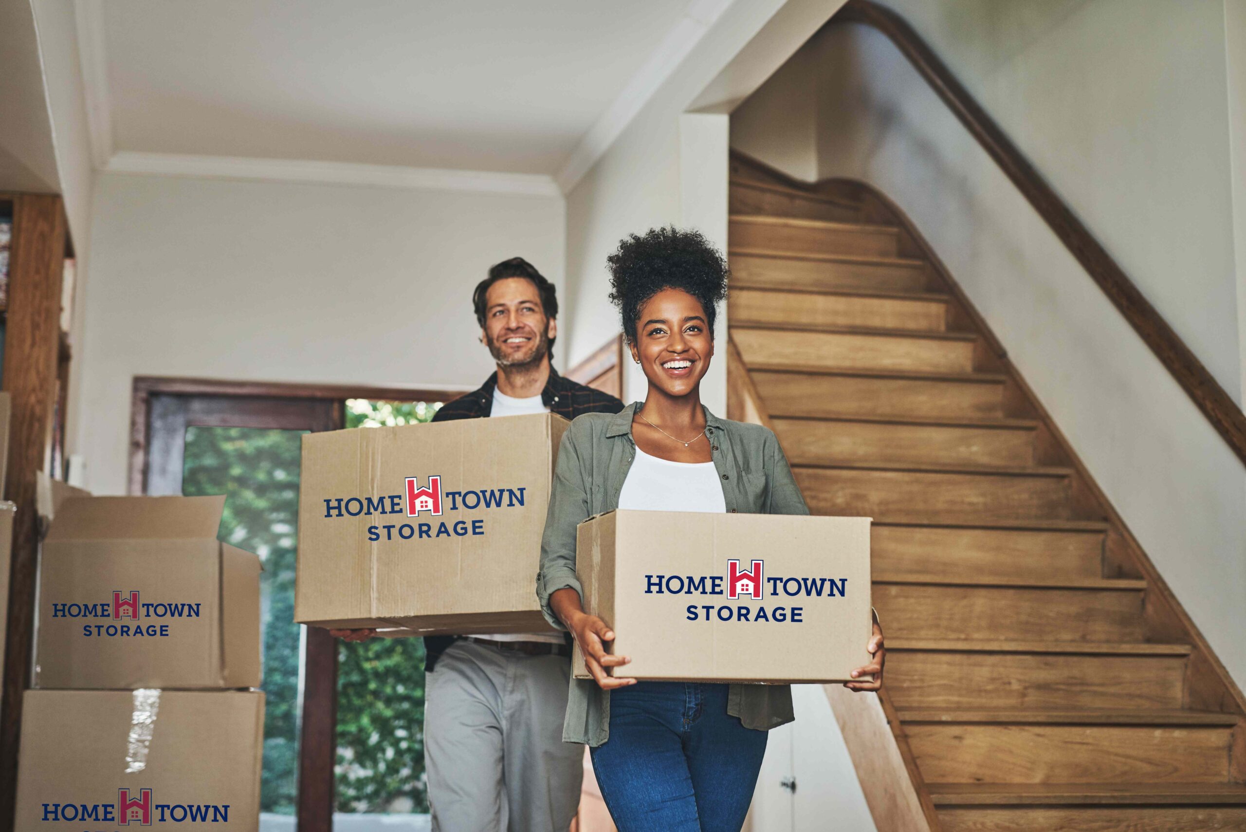 This image shows a smiling man and woman carrying moving boxes labeled "Home Town Storage"