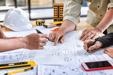 Three people review and discuss blueprints on a table covered with construction tools and a hard hat.