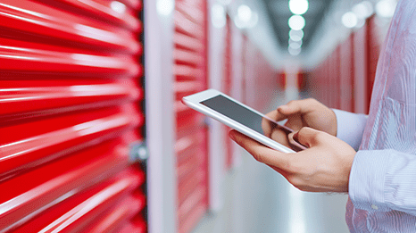A person holding a tablet stands in a hallway lined with bright red storage unit doors. The focus is on the hands using the tablet, suggesting inventory or management tasks.