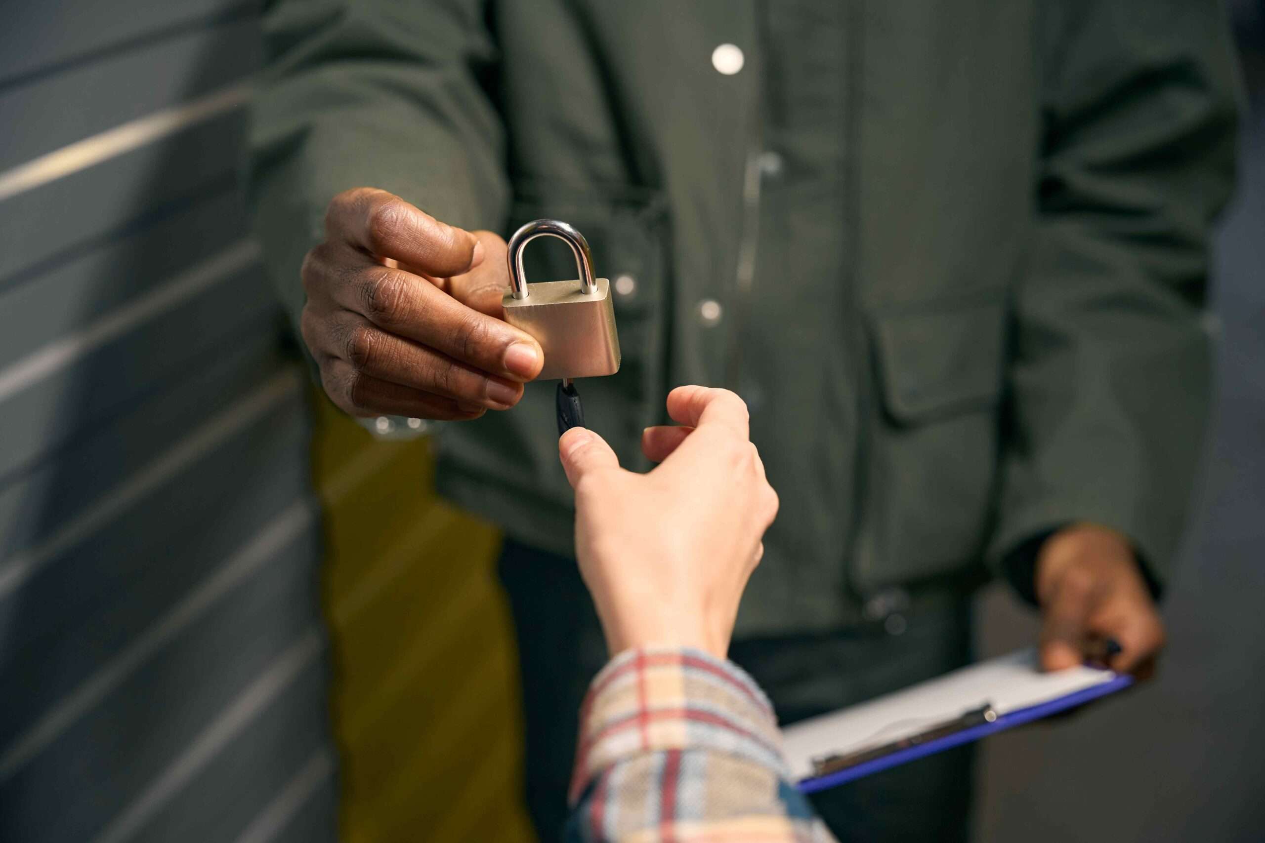 A person hands a padlock and key to another person in front of metal storage doors. The exchange suggests securing or accessing a storage unit.